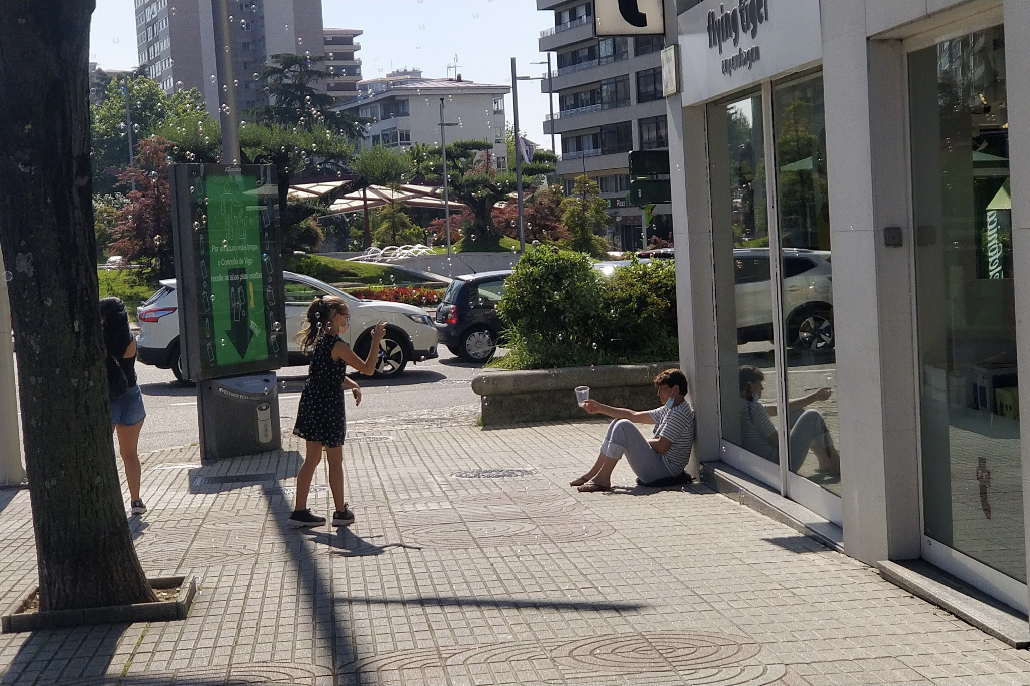 Una mujer pidiendo limosna mirando a una niña jugar con pompas de jabón